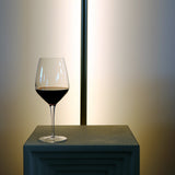Atelier Cabernet wine glass filled with red wine standing on a white surface and a long lamp in the background