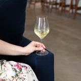 Hand holding Atelier Chianti wine glass filled with white wine on a dark blue velvet sofa