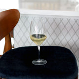 Atelier Chianti wine glass filled with white wine standing on a dark blue velvet chair with a white backsplash in the background