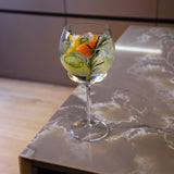 Backdoor '20s Gin & Tonic glass filled with a clear cocktail mixed with cucumbers, orange twist and rosemary standing on a marble surface and a neutral background