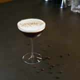 Backdoor '20s Hemmingway Special Cocktail Glass filled with Espresso Martini and garnished with coffee powder, standing on a brown surface decorated with whole coffee beans.