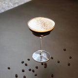Backdoor '20s Hemmingway Special Cocktail Glass filled with Espresso Martini and garnished with coffee powder, standing on a brown surface decorated with whole coffee beans.