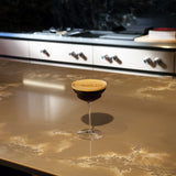 Backdoor '20s Hemmingway Special Cocktail Glass filled with Espresso Martini and garnished with coffee powder, standing on a brown marble surface and blurred kitchen in the background