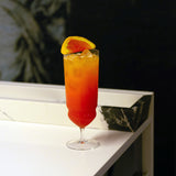Backdoor '20s Tequila Sunrise Cocktail Glass filled with a bright red cocktail and garnished with a slice of blood orange, standing on a white marble surface dark blurred background