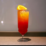 Backdoor '20s Tequila Sunrise Cocktail Glass filled with a bright red cocktail and garnished with a slice of blood orange, standing on a wooden surface and a neutral background