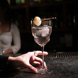 Hand holding a Backdoor '20s Barracuda Cocktail Glass filled with a clear cocktail and garnished with a Lychee on a bar surface and a dark background