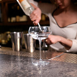 Empty Backdoor '20s Great Gatsby Cocktail Glass standing on a bar surface and a bartender making a cocktail in the background