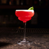 Bright pink cocktail in a Backdoor '20s Nick & Nora Cocktail Glass standing on a bar surface and a dark background