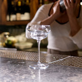 Bartender shacking a cocktail in the background of an empty Backdoor '20s Nick & Nora Cocktail Glass that is standing on a bar surfac