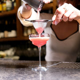 Hands pouring a pink cocktail into a Backdoor '20s Nick & Nora Cocktail Glass that is standing on a bar surface