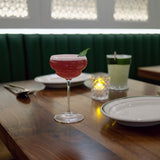Bright pink cocktail in a Backdoor '20s Nick & Nora Cocktail Glass standing on a restaurant table with a set table background