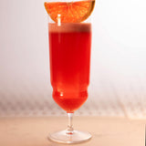 Backdoor '20s Tequila Sunrise Cocktail Glass filled with a bright red cocktail and garnished with a slice of blood orange, standing on a blurred light surface and a blurred background