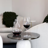 Magnifico decanter filled with red wine standing next to a Vinoteque Riserva wine glass filled with red wine on a silver side table, white chairs are in the background
