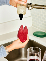 Person using a red kitchen tool to drain liquid from a bottle over a sink.