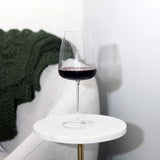 Talismano Italian Premium Bordeaux Red Wine Glass filled with a red wine standing on a white side table with a white chair and a blanket in the background