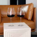 Two Talismano Italian Premium Bordeaux Red Wine Glasses filled with red wine standing on a white stool with a brown leather sofa in the background
