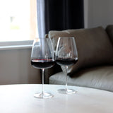 Two Talismano Bordeaux Red Wine Glass filled with a red wine standing on a white surface with a grey sofa in the background