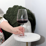 Hand holding a Talismano Italian Premium Bordeaux Red Wine Glass filled with a red wine standing on a white side table with a white chair and a blanket in the background