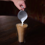 Thermic double-wall latte cup filled with latte and a hand pouring milk inside, standing on a dark wooden surface