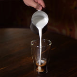 Thermic double-wall latte cup filled with latte and a hand pouring milk inside, standing on a dark wooden surface