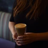 Two hands holding a Thermic double-wall latte cup filled with latte on a dark background