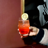 hand holding a Backdoor '20s Bramble glass filled with a red cocktail garnished with a slice of lemon on a dark red background with a framed black and white photo