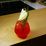 Atelier stemless glass filled with bright red cocktail and garnished with apple and thyme standing on a wooden surface