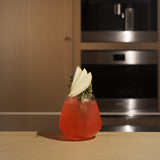 Atelier stemless glass filled with bright red cocktail and garnished with apple and thyme standing on a wooden surface and blurred kitchen appliances in the background