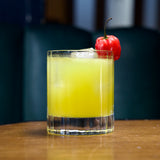 Bach DOF glass with a bright yellow cocktail garnished with a red habanero standing on a wooden surface and a drack background