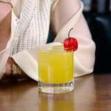 Bach DOF glass with a bright yellow cocktail garnished with a red habanero standing on a wooden surface an a person in ivory outfit in the background