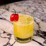 Bach DOF glass with a bright yellow cocktail garnished with a red habanero standing on a marble surface