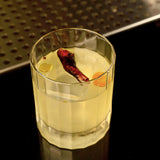Bach DOF glass with a light yellow cocktail garnished with a red dried hot pepper standing on a dark bar surface