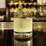 Bach DOF glass with a light yellow cocktail garnished with a red dried hot pepper standing on a dark bar with blurred bottles in the background