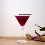 Bach Martini glass with a deep purple cocktail garnished with lavender standing on a wooden surface next to flowers and a white wall in the background