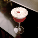 Bach Retro Fizz Cocktail glass with a red cocktail and white foam garnished with a raspberry standing on a dark surface