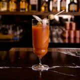 Red cocktail in a Backdoor '20s Tequila Sunrise glass with garnishes of olives on a bar counter, blurred bottles in the background