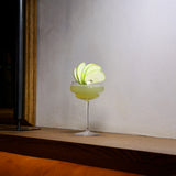 Backdoor '20s Great Gatsby Cocktail Glasses with a light green cocktail garnished with an apple standing on a wooden surface with a white wall and a picture frame in the background