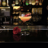 Backdoor '20s Great Gatsby glass with a orange cocktail garnished with a single red rose petal on a dark bar surface next to a red rose with blurred bottles in the background