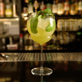 Backdoor '20s Gin & Tonic glass with a yellow cocktail garnished with mint leaves standing on a bar counted with blurred bottles in the background