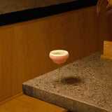 Backdoor '20s Italian Premium Nick & Nora Cocktail glass filled with a pink cocktail and garnished with line zest standing on a marble surface and a wooden bench in the background
