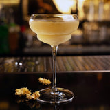 Backdoor '20s Nick & Nora Cocktail Glass filled with a clear light yellow cocktail standing on a dark bar surface with dry flowers next to the glass and blurred background
