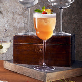Backdoor '20s Barracuda Cocktail Glass filled with a orange cocktail and garnished with mint, cherry and pineapple standing on a wooden tray with a old wooden box with cocktail glasses on top in the background