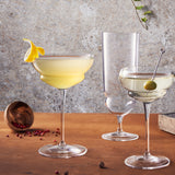 Backdoor '20s Hemmingway Special Cocktail Glass filled with a light yellow cocktail and garnished with a lemon twist standing on a wooden surface next to two other glasses from the collection