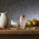 Two Backdoor '20s stemless glasses with cocktail and whisky on a wooden table decorated with a white vase and fruits