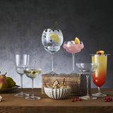 Six stemmed glasses of different shapes from the Backdoor '20s collection filled with cocktails and standing on a wooden table decorated with fruits.