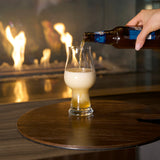 Hand pouring beer from a bottle into a Birrateque IPA Beer Glass standing on a wooden surface and a fireplace in the background