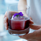 Two hands holding a Classico DOF Glass filled with a deep purple cocktail and big ice cube, garnished with a purple flower on a light background