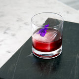 Classico DOF Glass filled with a deep purple cocktail and big ice cube, garnished with a purple flower on a light background