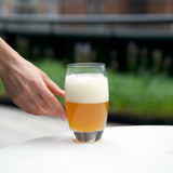 hand reaching a Crescendo Highball drinking glass half filled with beer and thick foam, standing on a white surface and blurred background