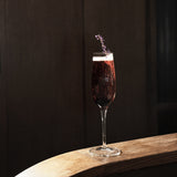 Crescendo Flute Glass filled with a deep purple cocktail garnished with lavender standing on a thin wooden surface and a dark background